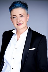 Judith with mauve hair looking at the camera wearing a white blouse and black jacket