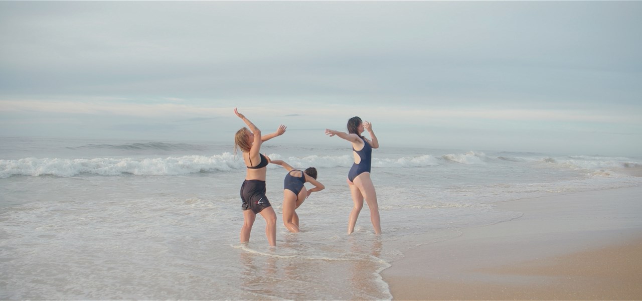 Three people dance on a beach in shallow water.