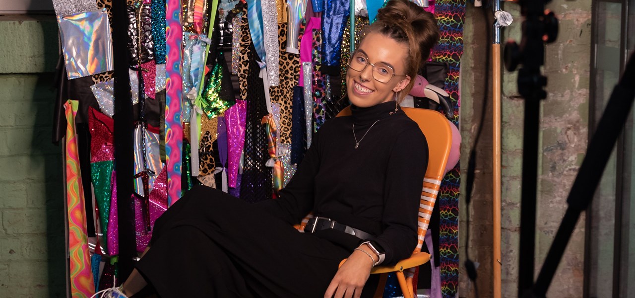 Person dressed in black clothes and white platform boots with a lot of brightly coloured clothing hanging behind them