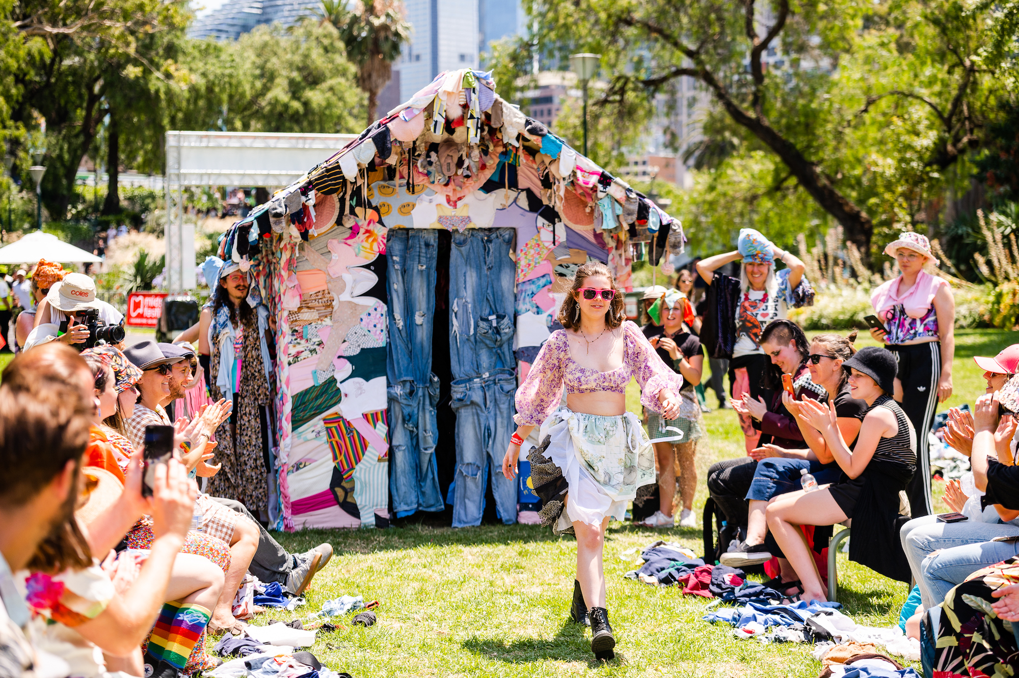 A colourfully dressed person walking a lawn catwalk in a park with a colourfully decorated shed in the background