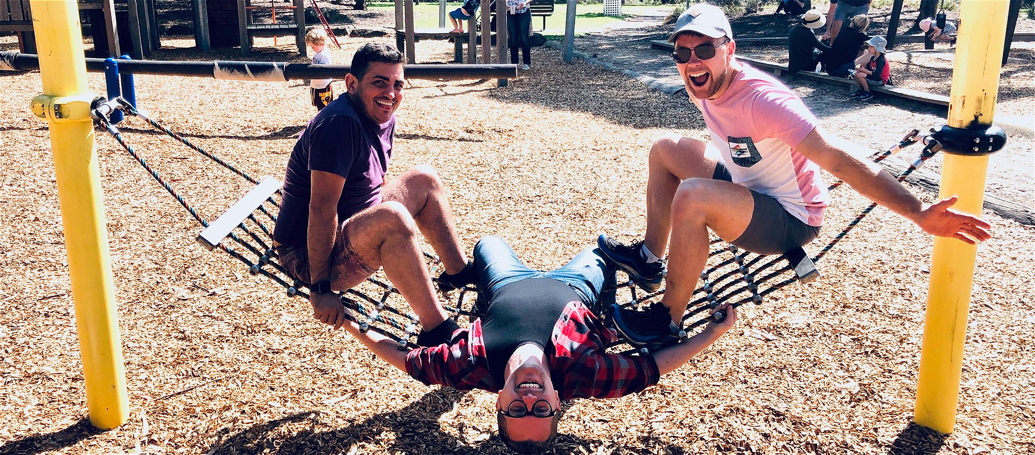 Three people on a net hammock in the playground, smiling at the camera.