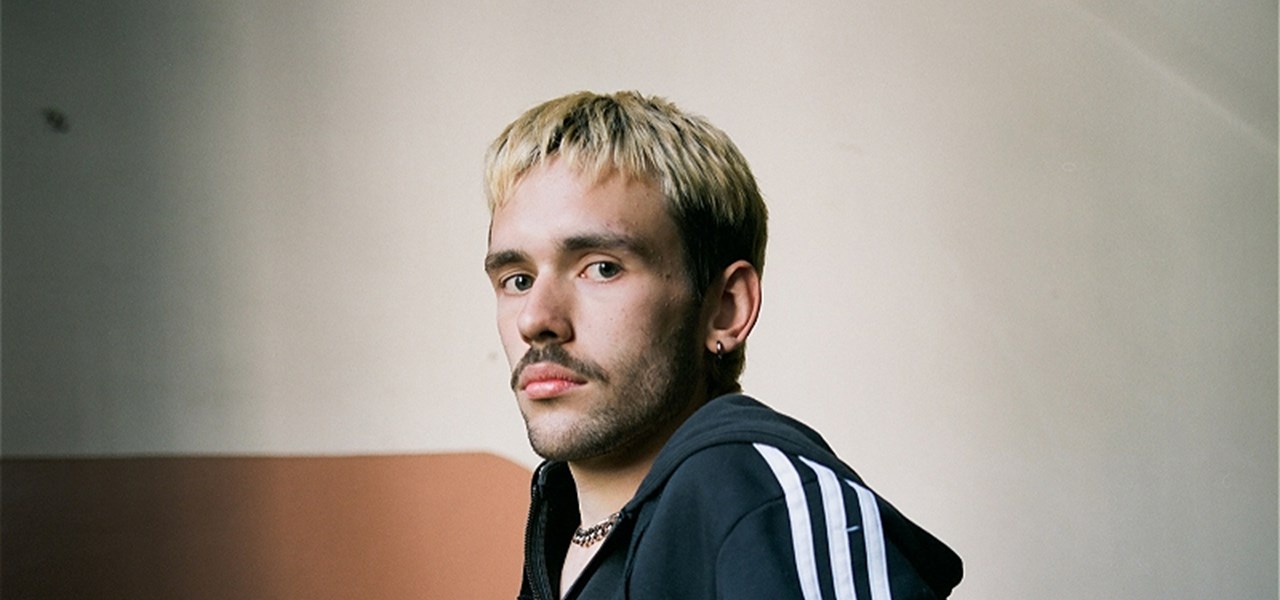 Head and torso of a dyed-blond male-identifying person facing the camera, wearing a tracksuit top. They have a moustache and beard stubble.