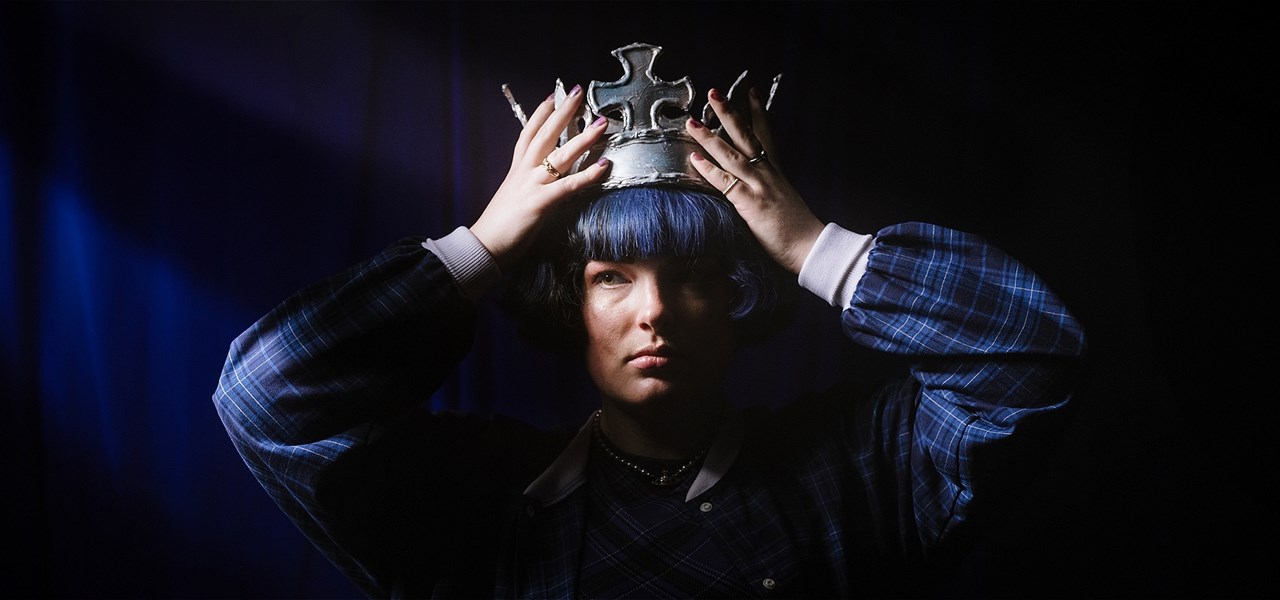 A person with a cheek length blue bob and their face split down the middle with pink and blue light wears a gold crown while staring into the camera.