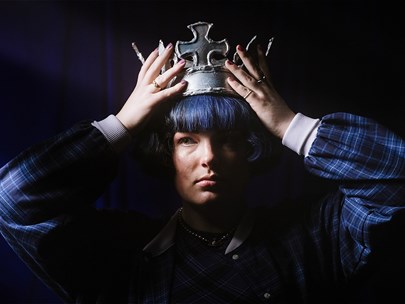 A person with a cheek length blue bob and their face split down the middle with pink and blue light wears a gold crown while staring into the camera.