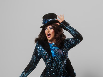 Valerie Hex in a dark blue, glittery suit wearing a top hat, with a grey background