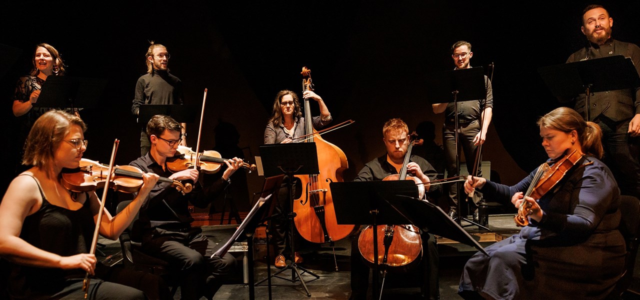 A group of musicians, some playing string instruments and some singing, all dressed in formal black attire.