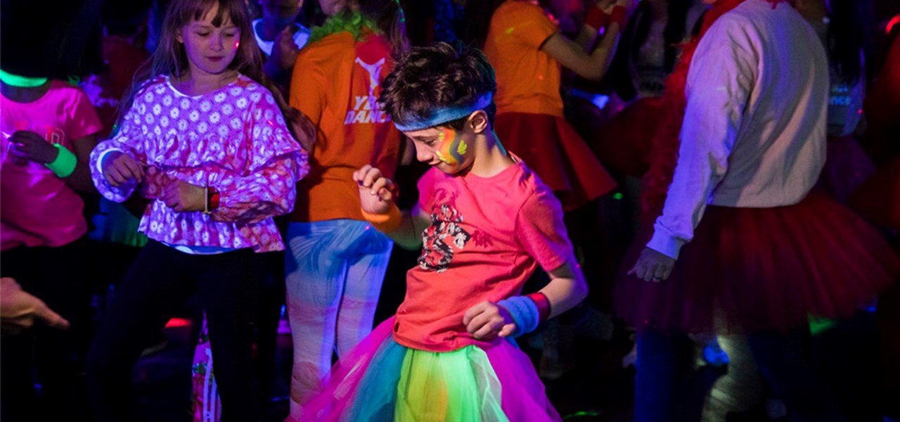 Kids dressed in glittery colours dancing. The kid in the foreground is wearing a rainbow dress
