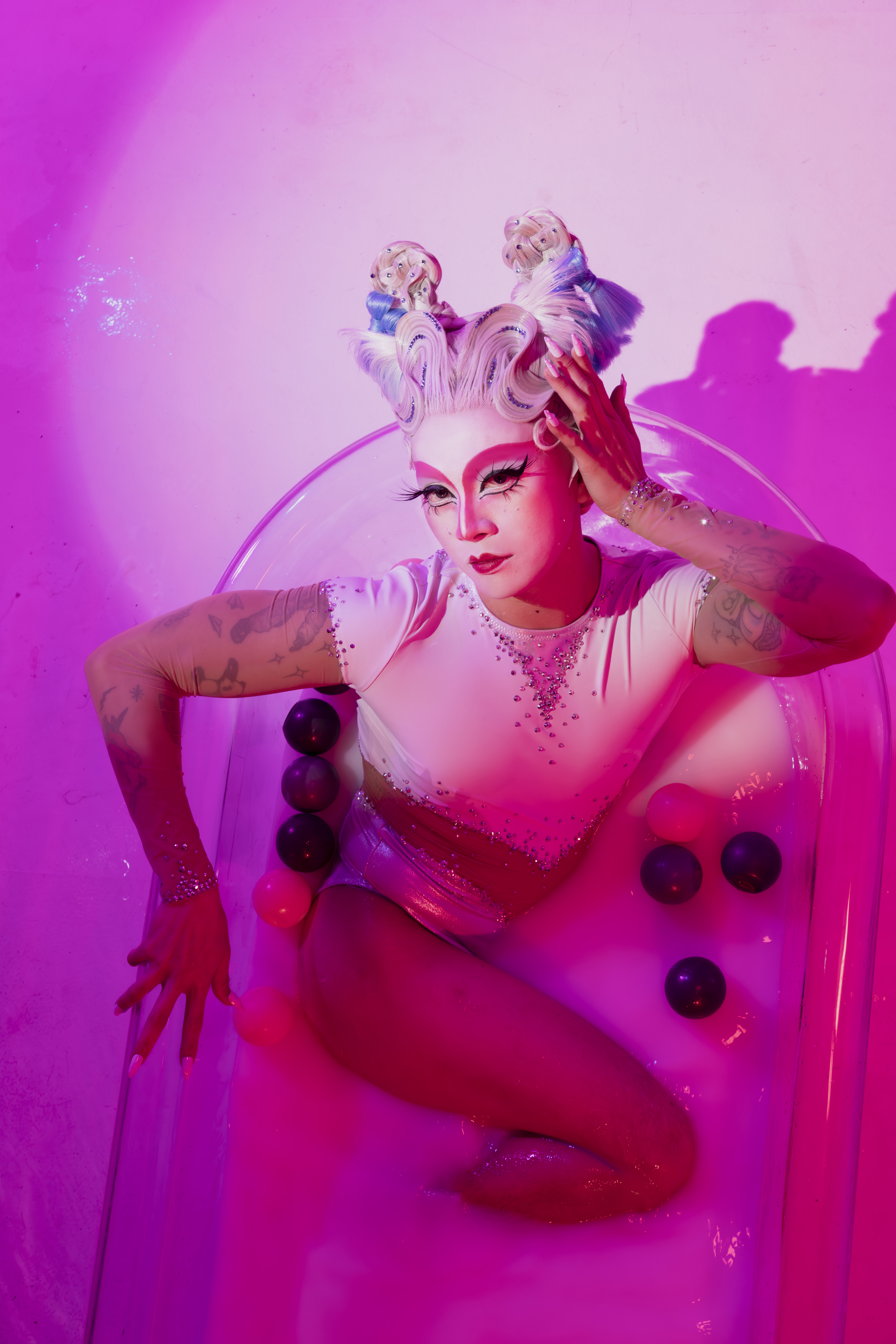 A fully clothed person is sitting in a bubble bath. The lighting has pink tones. 