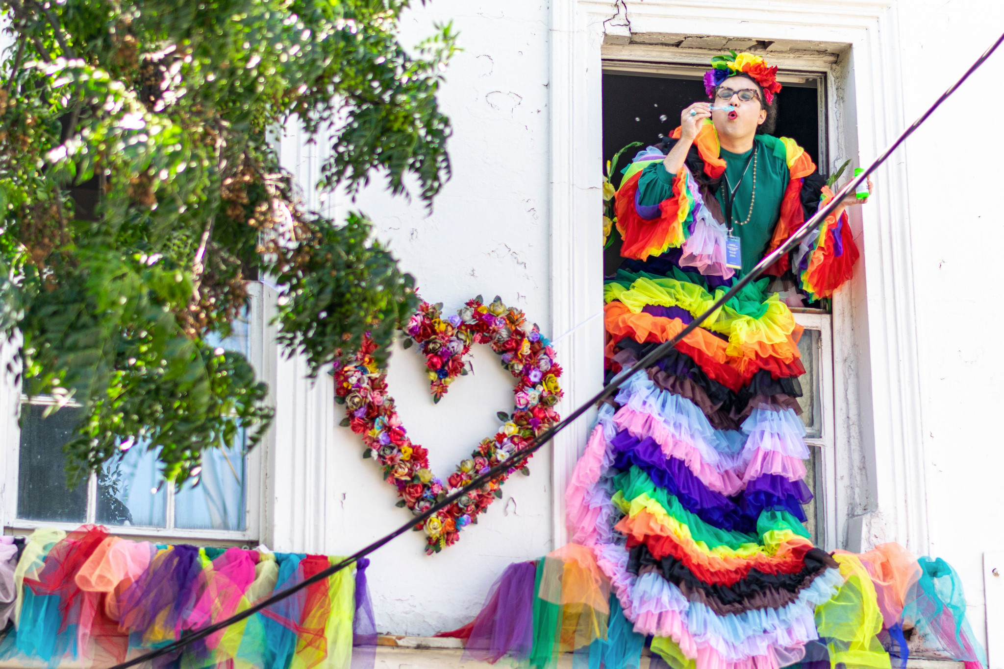 Mama Alto hanging out of a window wearing a pride-colours dress, blowing bubbles