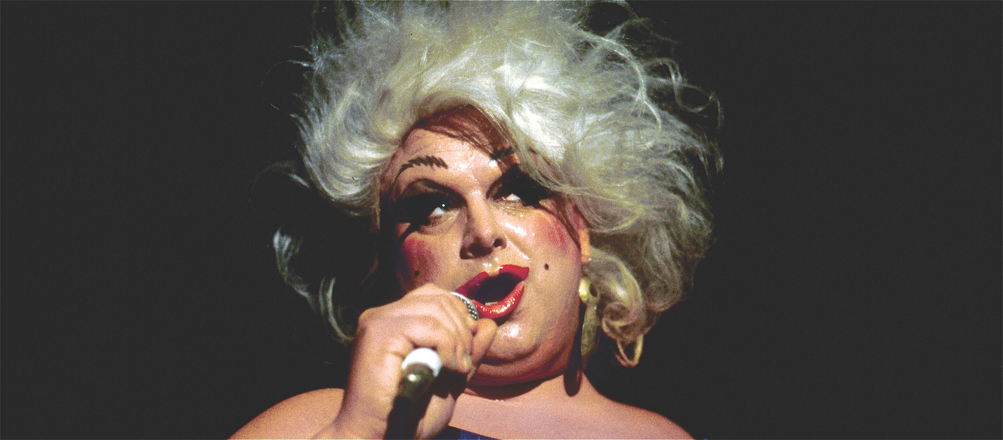 Image of the iconic drag queen Divine on the microphone in a blonde wig