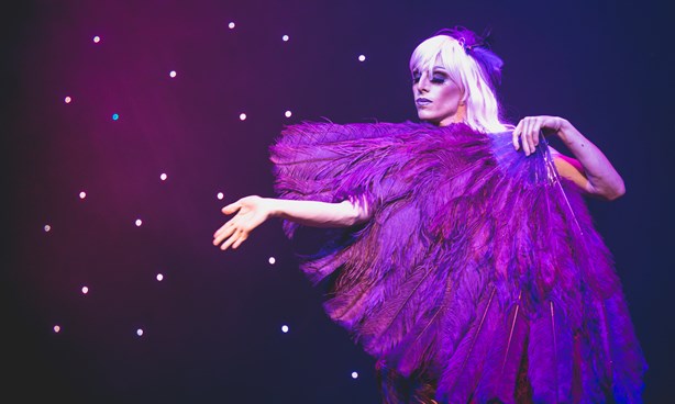 A performer in a "feather-clad fairy" outfit, one hand reaching out; mauve lighting with stars behind