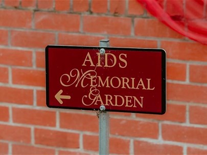 A small maroon plaque with a black boarder that reads -AIDS Memorial Garden- in gold font and an arrow pointing left.