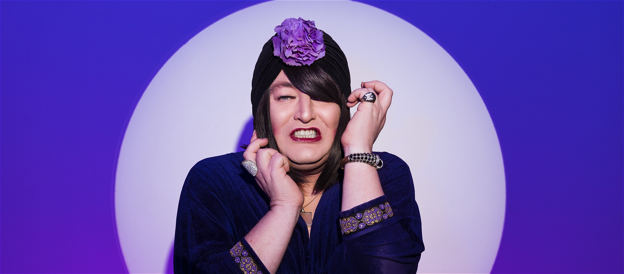 Tina wears a turban and has her hands up near her face with a spotlight and purple background