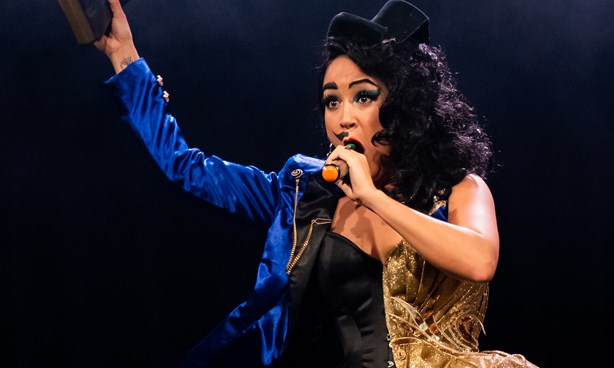 Person dressed in black and blue on the left, gold on the right, wearing a top hat, holding a book and speaking into a microphone