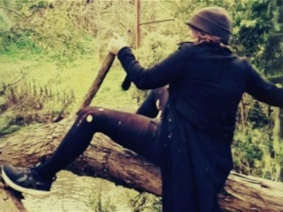 A warm image of a woman in a flowing jacket riding a broken tree like a cowboy in nature.