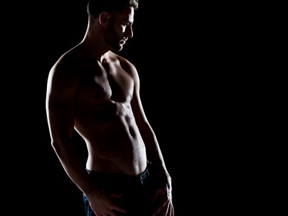 A muscular shirtless man stands, dimly lit, facing away from the camera.