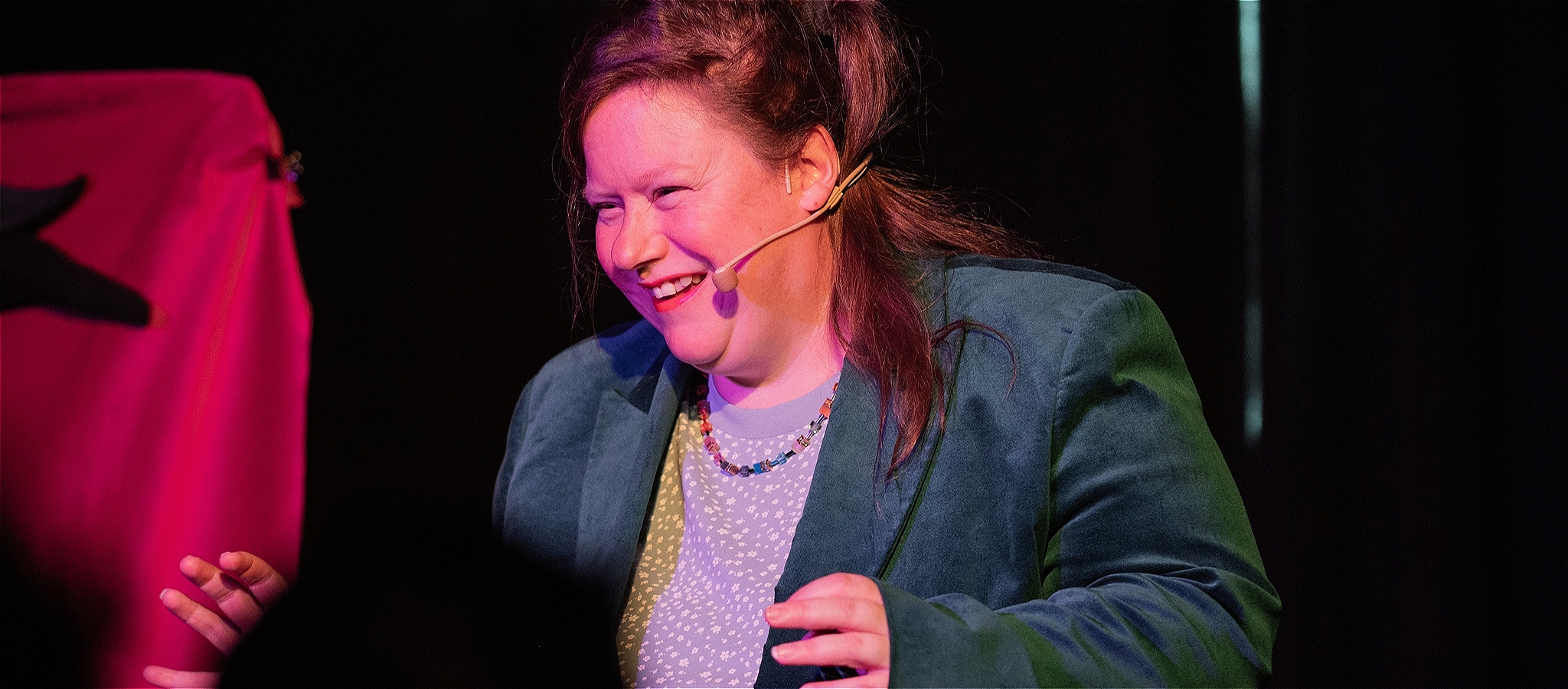 A woman, smiling, on stage, wearing a dark green blazer, blue tshirt and rainbow necklace. With a microphone headset.