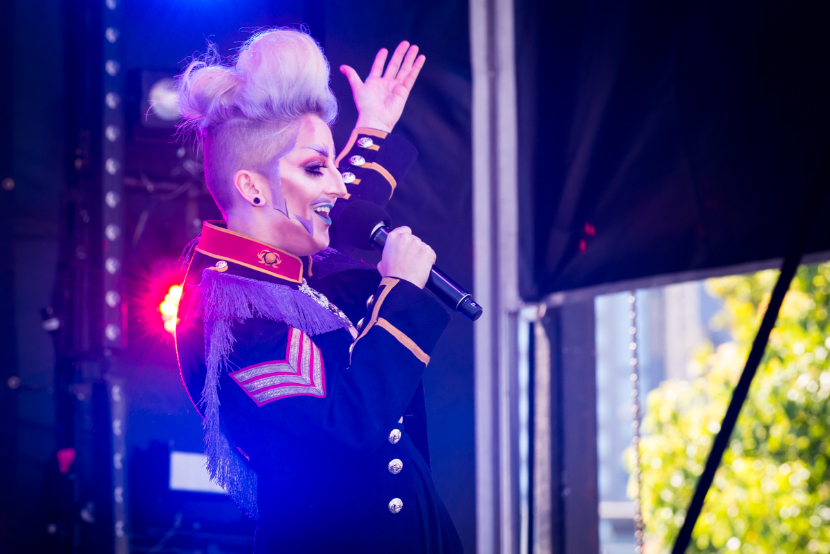 Drag king on stage with mic in one hand and the other hand raised. They are wearing a military style outfit.