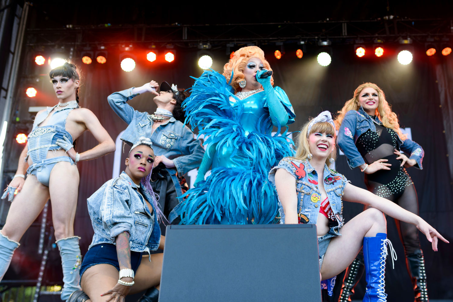 Drag queen singing, surrounded by elegantly dressed show people; all costumes are blue 