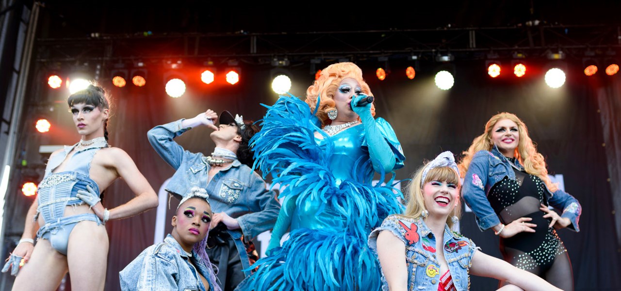 Drag queen singing, surrounded by elegantly dressed show people; all costumes are blue 