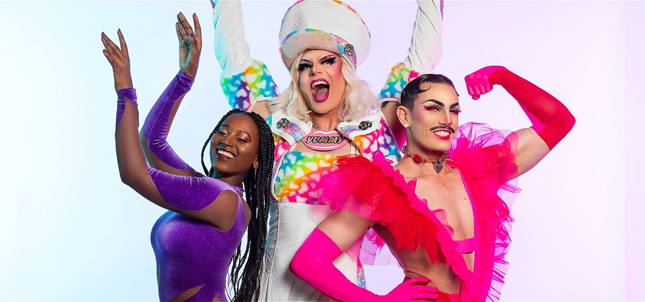Three performers from YUMMY pose with their hands up. Soliana is in purple, Valerie Hex is in rainbow, and Jarred is pink and red.
