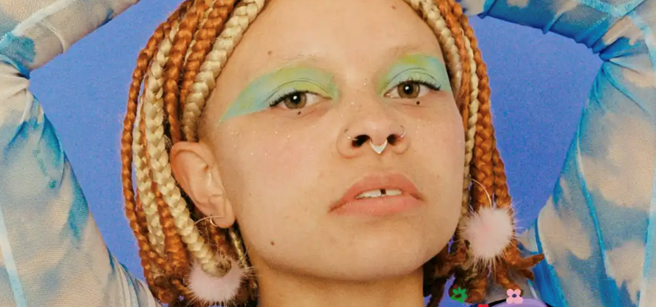 Young person with colourfully plaited hair holding their arms above their head. 