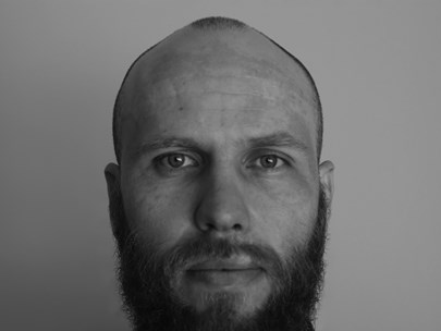 Black and white headshot of Sam looking serious, sporting a full beard and moustache