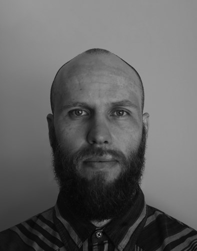 Black and white headshot of Sam looking serious, sporting a full beard and moustache