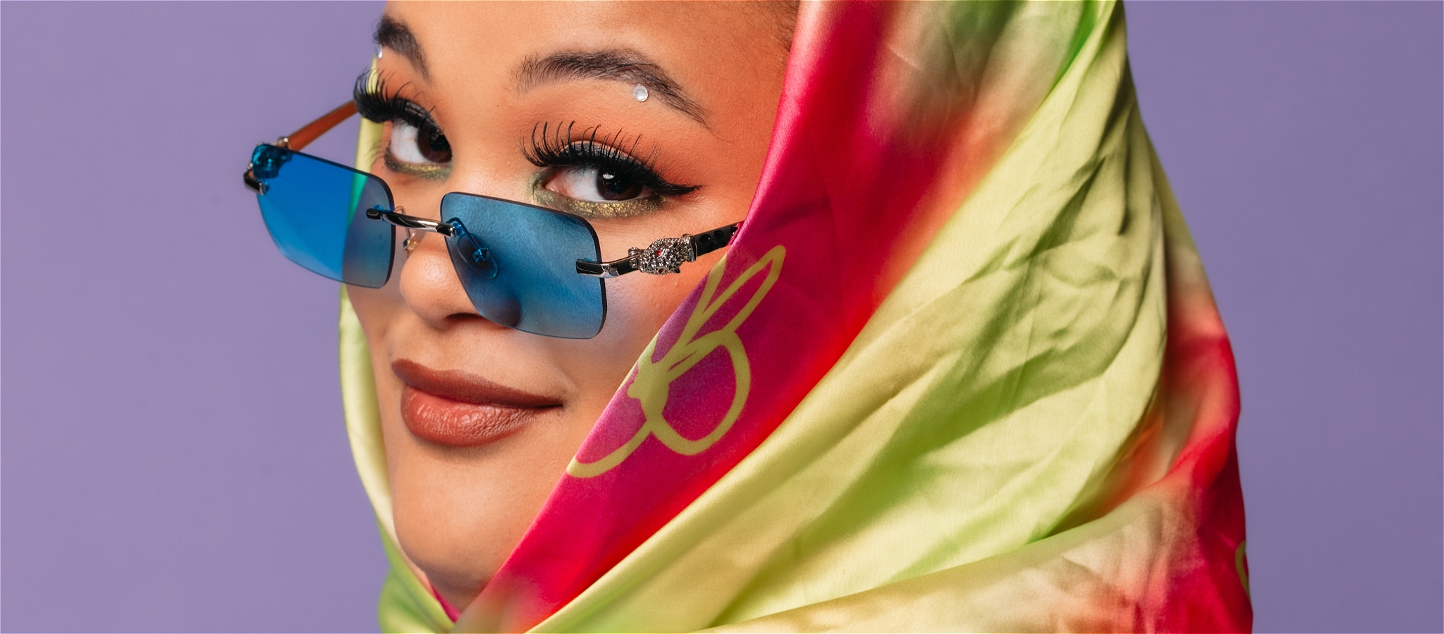 Close-up of Milo Hartill with braided hair, a vibrant headscarf, and blue sunglasses, posing in front of a purple backdrop.