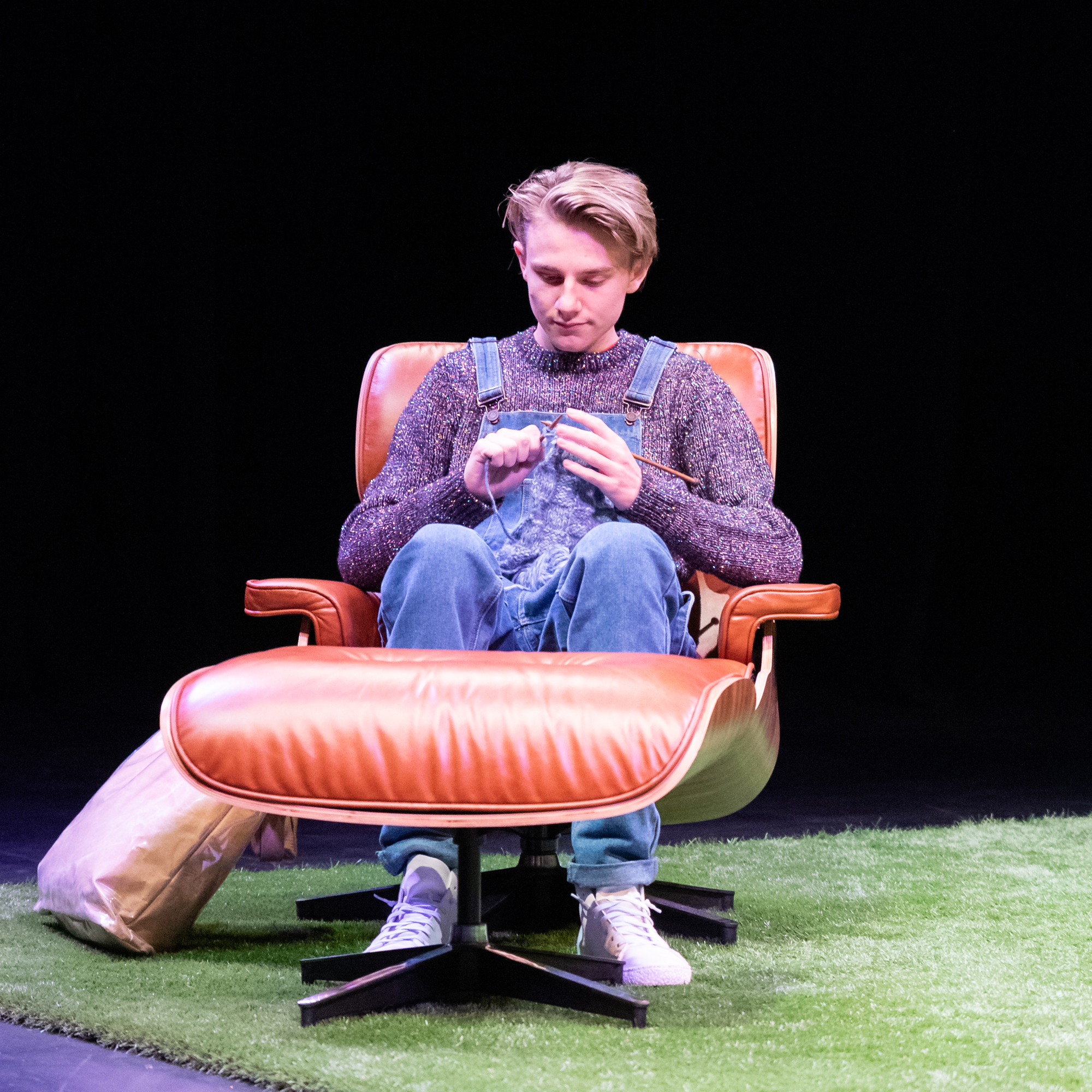 Man dressed in overalls and knitted jumper sitting in leather chair, knitting