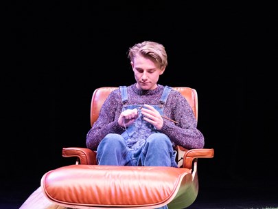 Man dressed in overalls and knitted jumper sitting in leather chair, knitting