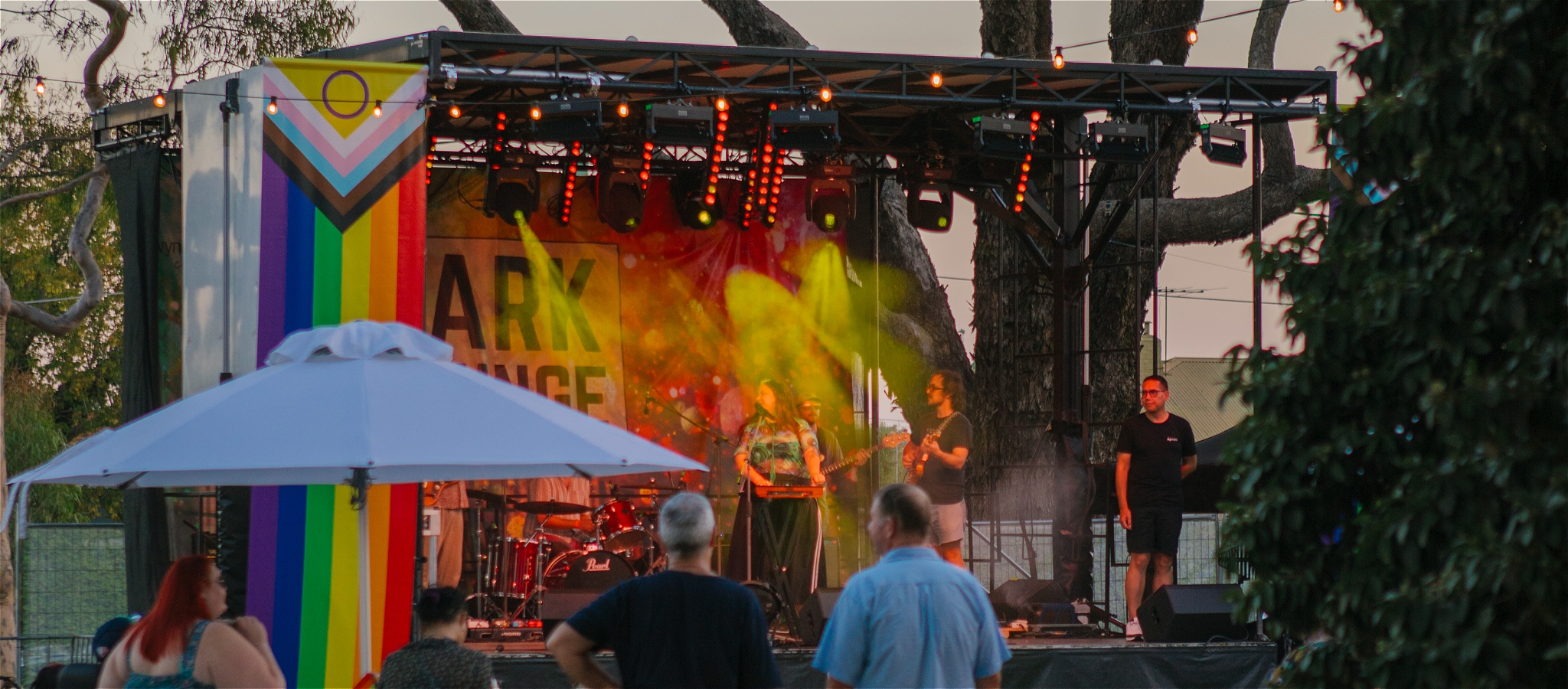 People Gather around a large umbrella as band performs on the Park lounge stage at Dusk