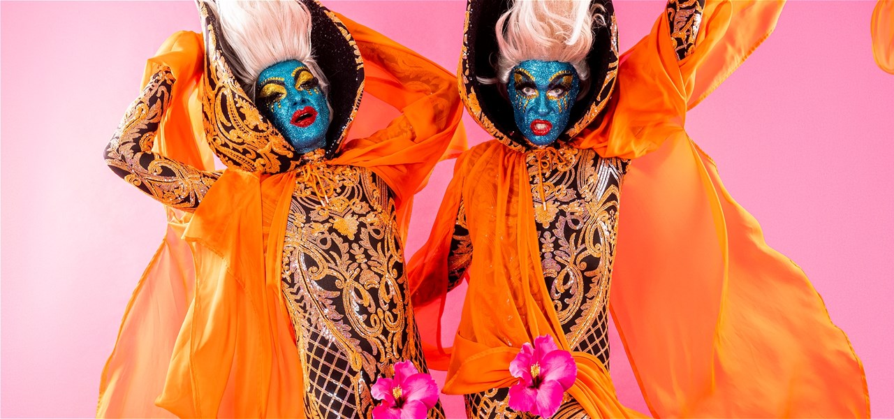 Two performers stand in front of a pink background wearing windblown orange capes and hibiscus flowers, glare dynamically at the camera