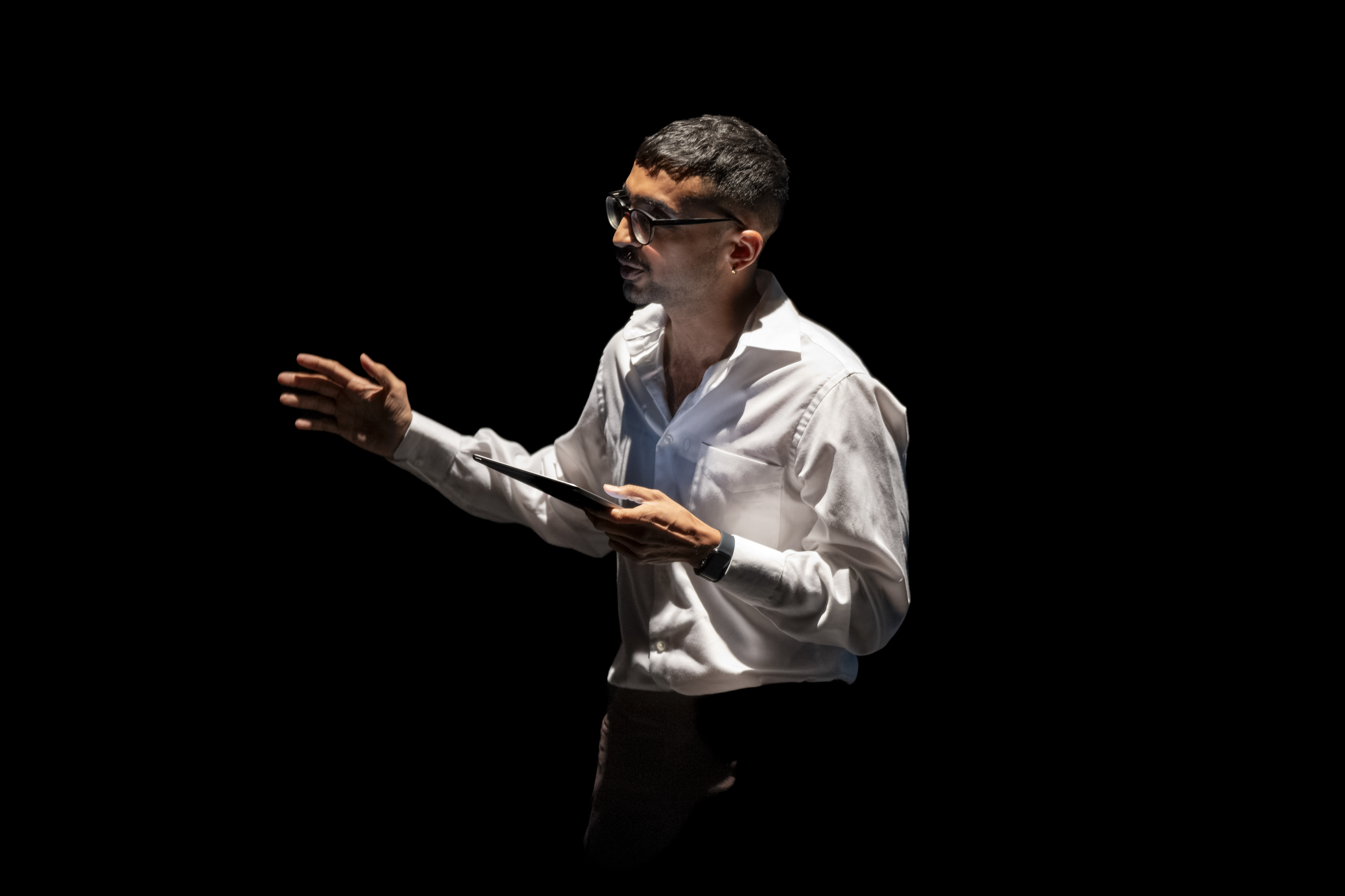 Danish Sheikh is lit in white light against a black background. Wearing a white shit, black trousers and glasses and reading an extract from their play from an iPad.