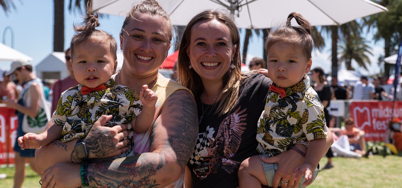 Two women at Midsumma Carnival, standing close to each other, each holding a young child