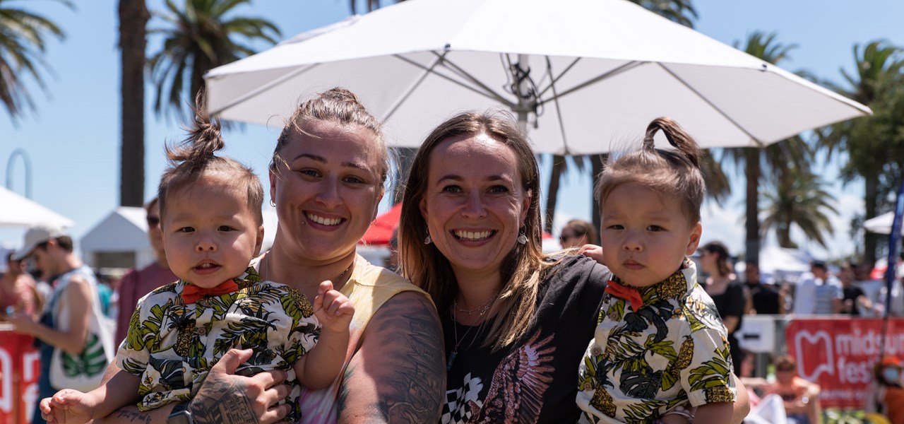 Two women at Midsumma Carnival, standing close to each other, each holding a young child