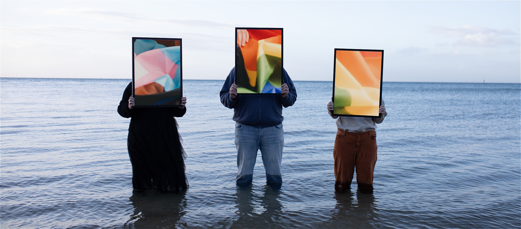 Three people standing in a calm ocean up to their knees and horizon line at their backs. Each holds a picture frame with imagery of the pride flag.