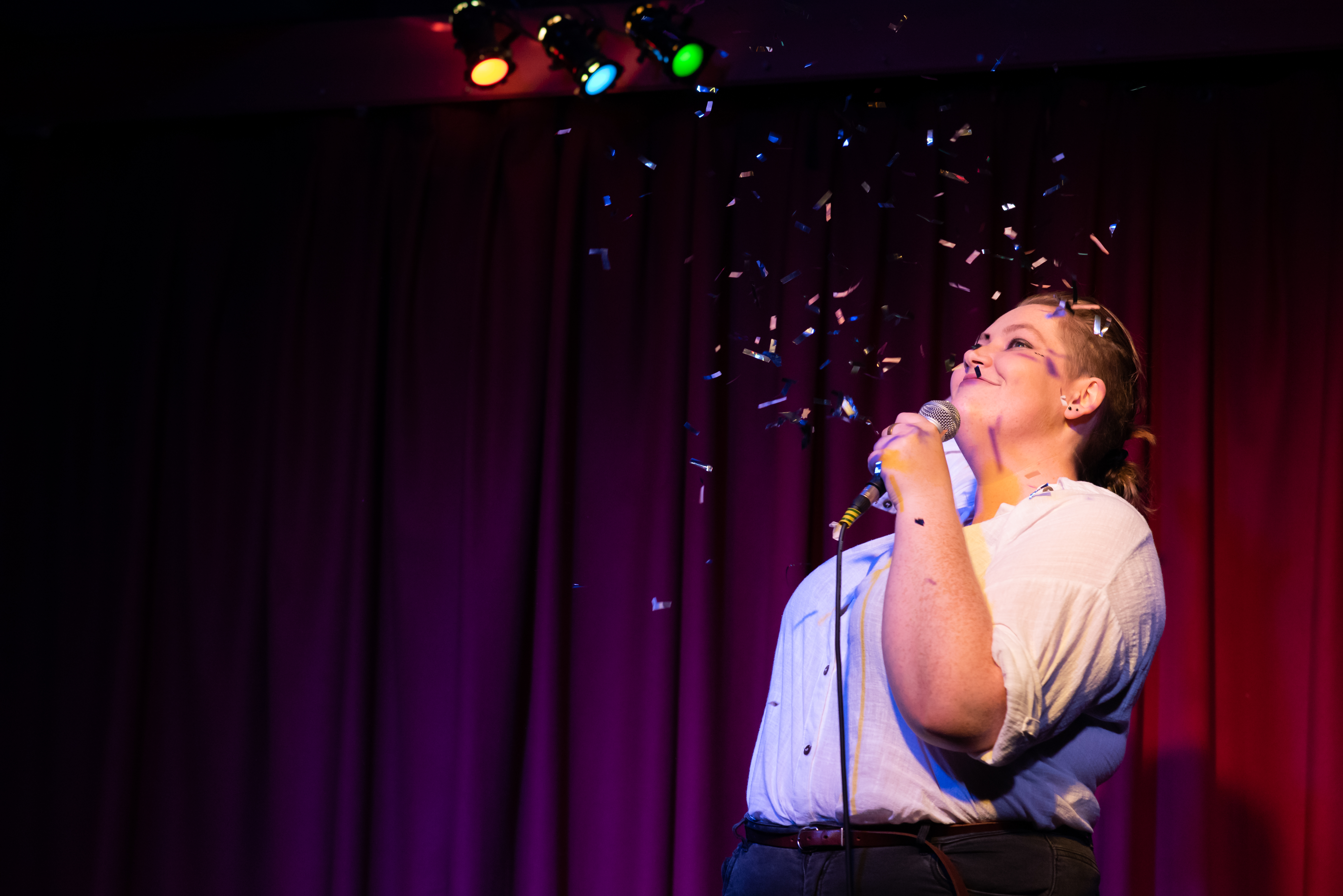 'Therapy is Expensive' performer on stage with glitter dropping onto them. Colour floodlights shining down above them.