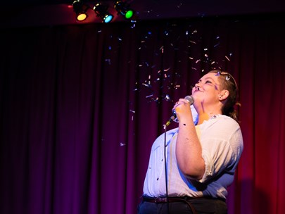 'Therapy is Expensive' performer on stage with glitter dropping onto them. Colour floodlights shining down above them.