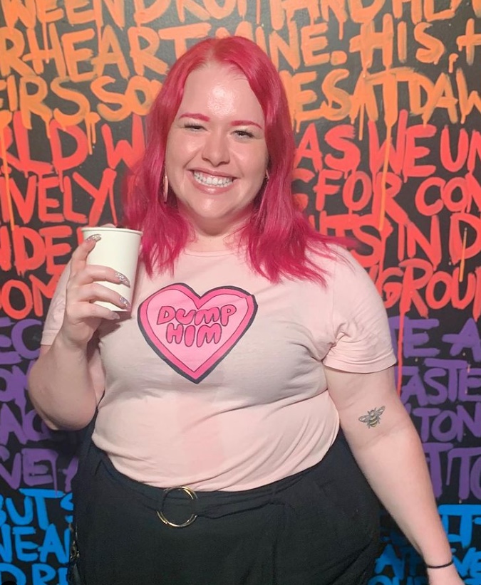 Emily Rose smiling broadly at the camera, holding a drink, wearing a "dump him" t-shirt