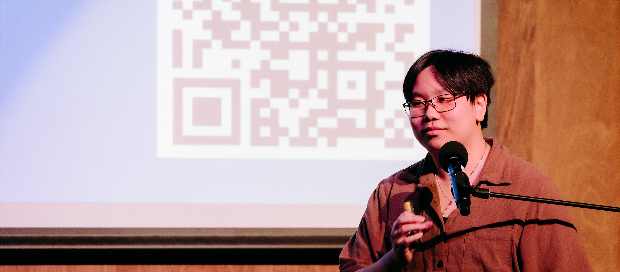 A person with dark hair and glasses standing in front of a microphone