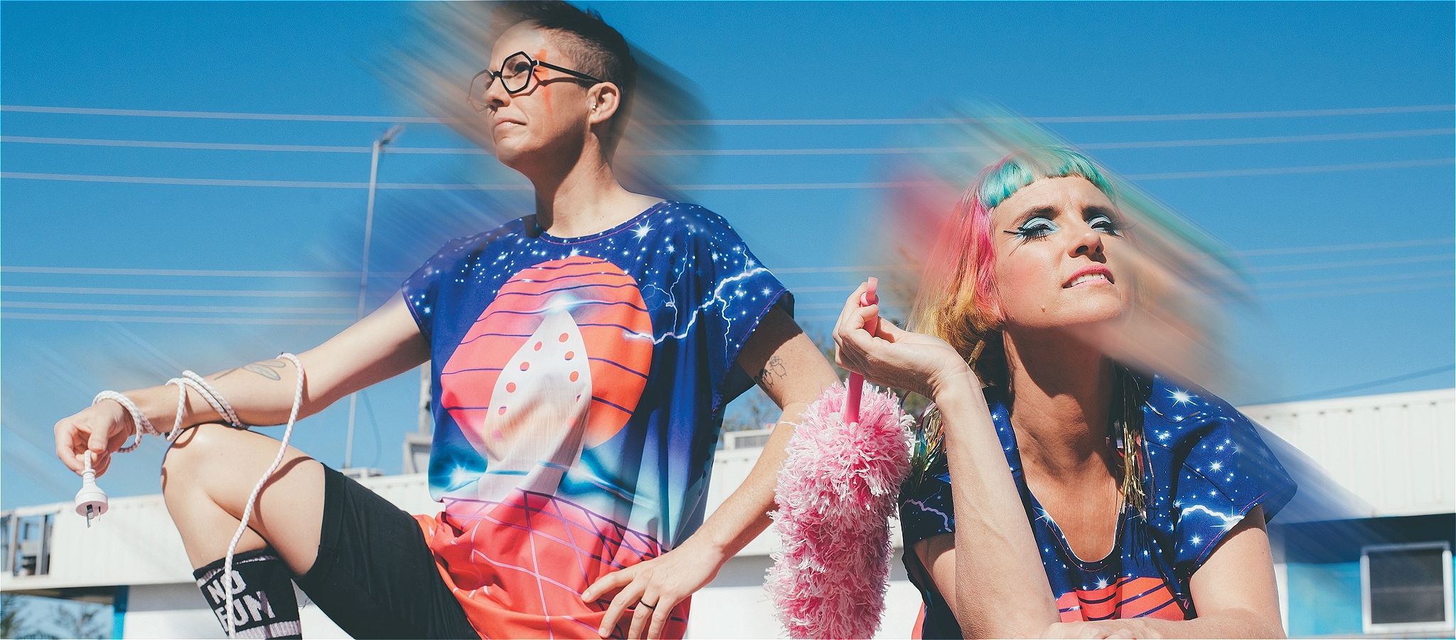A person with short brown hair and glasses holding an extension cord, and another person with rainbow coloured hair holding a pink feather duster