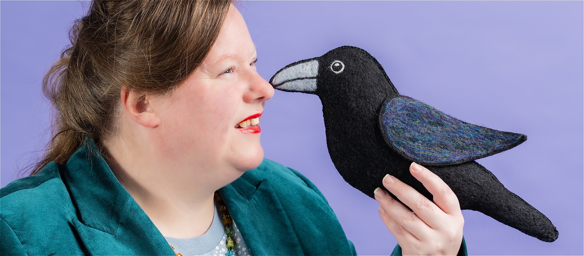 A smiling woman, in profile, wearing a dark green blazer, blue tshirt and rainbow necklace. Holding a plush raven.