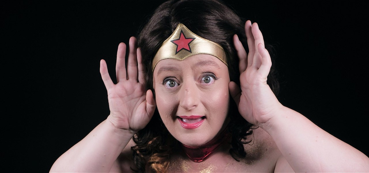 Woman looks directly into the camera with her hands by her ears. She is looking surprised and wearing a wonder woman gold headband with a star on it.