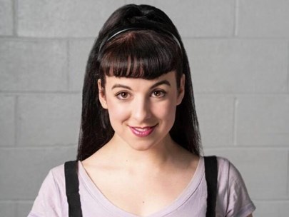 Rosanna has their long, straight hair in a high pony tail. They have a fringe which has been blow waves and wear a black headband behind it. They have pale skin and are wearing a white tshirt with black braces.