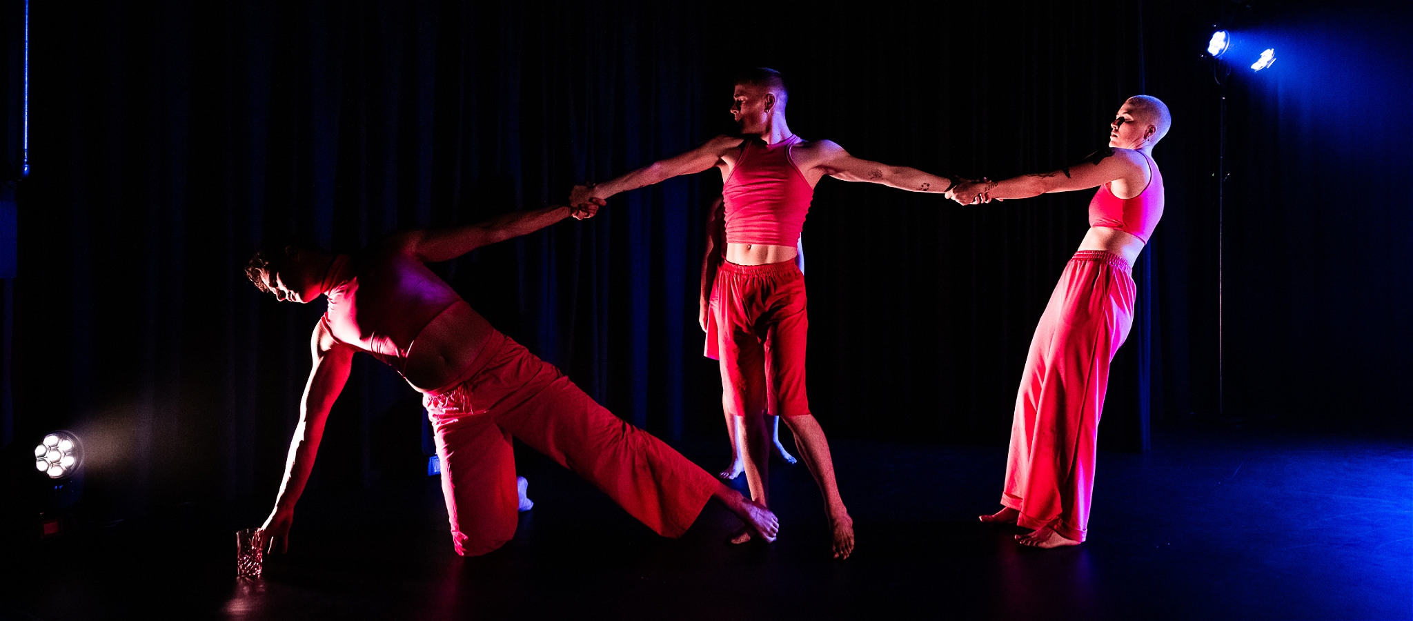 Three dancers hold each other by the hand while stretching across the stage.