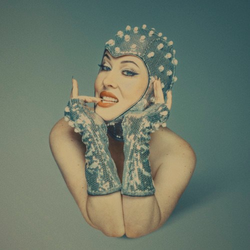 Tanzer wearing a slinky fitted bonnet and matching gloves holding their head between their hands with one finger touching their teeth.
