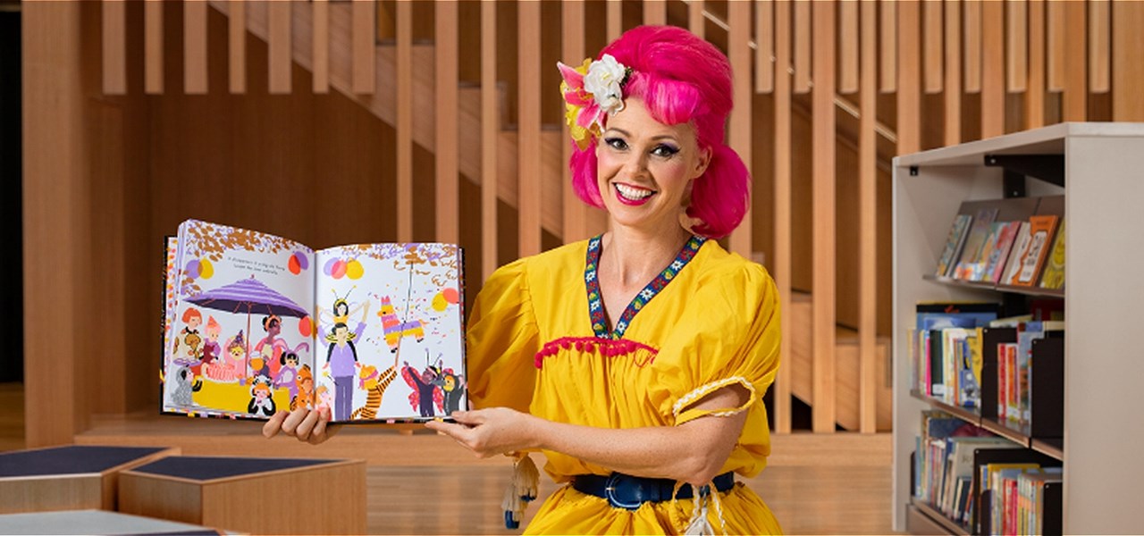 Drag queen dressed in yellow with red hair holding up a children's story book