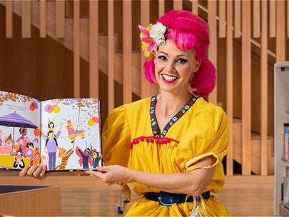 Drag queen dressed in yellow with red hair holding up a children's story book