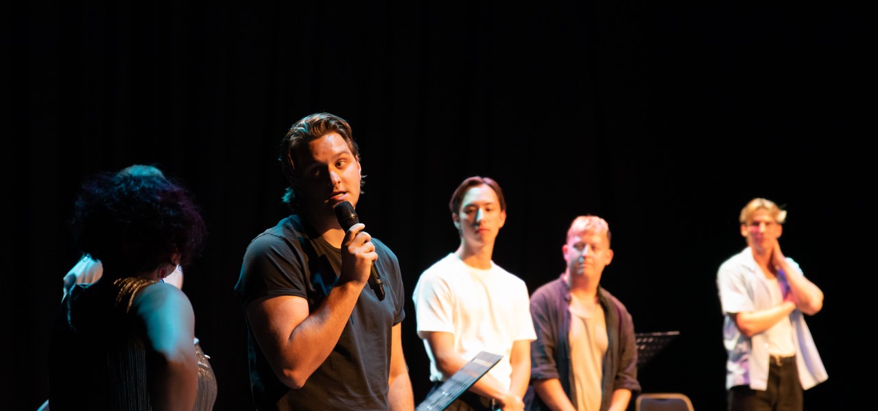 The playwright speaking into a microphone on stage while facing the compere (who has their back to us), watched on by the cast members